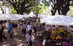 Foto de feiras com flores diversas em um ambiente muito arborizado, com várias pessoas passeando e realizando suas compras.