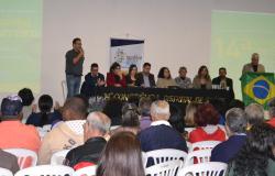 Mesa com autoridades e representantes de usuários, diante de auditório repleto de profissionais e usuáios do SUS.