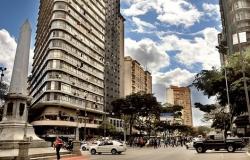 Praça Sete, no centro de Belo Horizonte, com um obelisco à esquerda e edifícios altos, rua e movimento pessoas e trânsito em um dia claro com nuvens.