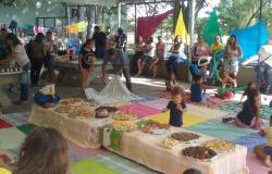 Mais de 30 pessoas, entre adultos e crianças, confraternizam em quadra coberta com mesas de quitures, e enfeites.