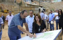 Prefeito Alexandre Kalil vê planta do Aglomerado Santa Lúcia, acompanhado por engenheira. Logo atrás, grupo de pessoas está reunido. Ao fundo, o casas e barracos do Aglomerado Santa Lúcia.
