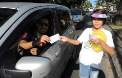 Uma criança segurando um balão amarelo e entregando um panfleto a motorista homem, com óculos escuros, no sinal.