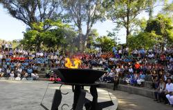 A foto mostra pessoas sentadas em arquibancadas de cimento, em um parque com árvores ao fundo. O céu está azul. A maior parte do público é constituída de alunos uniformizados. Em um plano superior, ao meio, está situada uma grande o pira em metal preto, e o fogo está aceso, uma chama grande e amarela.