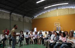 Vinte e oito pessoas em uma quadra coberta sentadas em cadeiras de plásticos brancas, segurando cadernos, olhando para um homem que está mediando  uma discussão sobre os eixos que estão sendo discutidos em uma pré-conferência