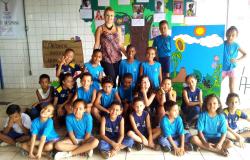 Crianças, estudantes da rede municipal de ensino, posam para a foto junto com professora. Ao fundo, painel de árvore.