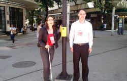 Dois Deficientes visuais aguardam ao lado do semáforo sonoro a hora de atravessar a rua.