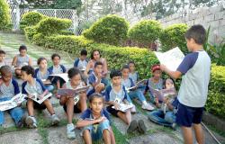 Crianças, estudantes da rede municipal de ensino, observam, sentados, o colega que lê de pé. Ao fundo, o jardim da escola.