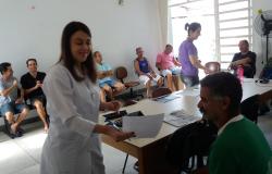 No primeiro plano, funcionária da saúde segura folha de exames de paciente. Ao fundo, fila com outros pacientes que aguardam atendimento. Todos são homens.
