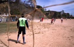 Campo de várzea. Goleiro está de costas. No campo, atletas disputam a bola