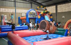 Cerca de 20 crianças se divertindo em uma quadra de esportes, com dois colchões infláveis com touros mecânicos. No da frente, uma menina está sentada em um touro de brinquedo marrom, no outro, um menino está de pé em cima do brinquedo. Há outro brinquedo chamado play gol e as crianças estão espalhadas pela quadra brincando.