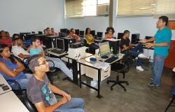 Cerca de nove jovens sentados em sala com mesas e computadores e Instrutor de pé. Foto ilustrativa