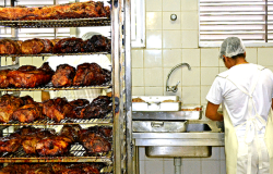 Mais de vinte frangos assados em prateleiras de metal na cozinha de restaurante popular; à direita, três formas retangulares de metal com frangos assados; mais à direita, funcionário corta frango assado em tábua de cozinha.