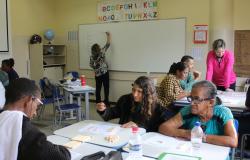 Professora escreve em quadro branco com mais de seis alunos adultos em carteiras à frente.