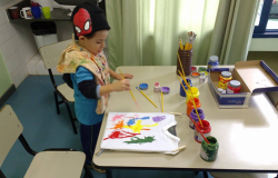 Criança com toca do Homem aranha pinta camiseta branca em mesa; à sua frente, vários potes de tinta.