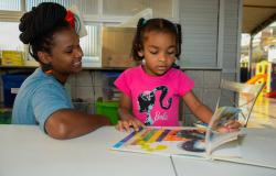 Mulher acompanha criança na leitura de livro infantil.