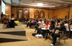 mais de cem professores e convidados em um auditório grande. No palco, vê-se uma mesa composta por pessoas da área de Educação, um telão e a secretária municipal de Educação, Ângela Dalben, com microfone.
