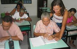 Idosos em sala de aula. Professora, em pé, auxiliando uma aluna.