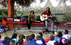 Mulher faz apresentação musical de violão para mais  de vinte crianças, ao fundo, outra mulher observa.