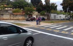 Mulher, de mão dada com criança, atravessa faixa de pedestre em rua de bairro. 