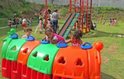 Cerca de quinze crianças, acompanhadas por educadores infantis, em pátio de Umei; uma brincam em túnel colorido, outras em escorregador e outras em cadeiras de balanço.
