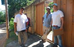 Dois agentes comunitários de saúde no portão de uma residência, junto ao proprietário, durante o dia. 