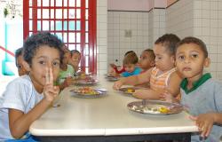 Cerca de oito crianças da Rede Municipal de Educação sentadas em uma mesa comendo merenda em pratos.