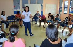 Cerca de treze jovens, sentados e em círculo, assistem a aula de instrutora que está de pé, em uma sala, durante o dia.