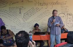 Homem declama poesia à frente de um mural com vários escritos. 