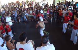 Mais de cinquenta pessoas assistem a jogo de capoeira em local coberto.
