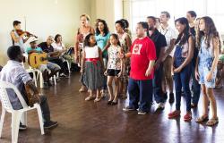 Cerca de doze alunos cantam de pé, em frente a orientador, que está sentado; ao lado, três pessoas com instrumentos musicais acompanham.
