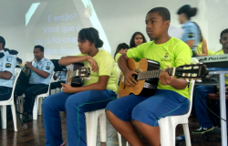 Duas crianças alunas de escola municipal tocam violão; ao lado, guardas municipais tocam outros instrumentos.