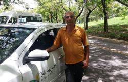 Taxista ao lado de táxi em rua arborizada, durante o dia.
