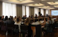 Mais de trinta pessoas em mesa em forma de "u" coma apresentação em powerpoint ao fundo com o título: Envelhecimento no século XXI.