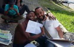 Morador em situação de rua abraça servidor e mostra desenho em folha branca. Os dois estão sentados no chão