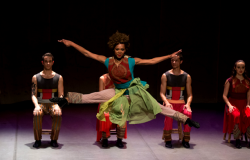 Mulher dá um pulo de dança em apresentação cênica, atrás, quatro pessoas estão sentadas.