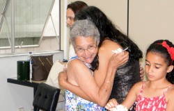Duas mulheres se abraçam, uma menina está mais à frente e à direita e uma mulher, atrás e à esquerda.