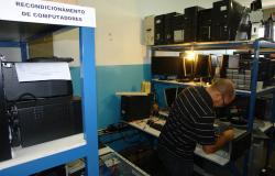 Técnico recupera computadores em sala com diversos componentes. Em uma estante à frente à esquerda, há uma placa com os dizeres: Recondicionamento de computadores. 