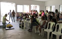Instrutor, ajoelhado perante voluntário, demonstra como fazer massagem cardiorrespiratória; em auditório, mais de vinte pessoas, sentadas ou de pé, acompanham a explicação.