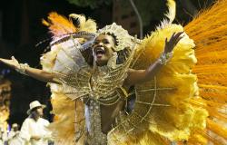 Passista de escola de samba, com adereços dourados, amarelos e laranjas, desfila no chão, à noite. 